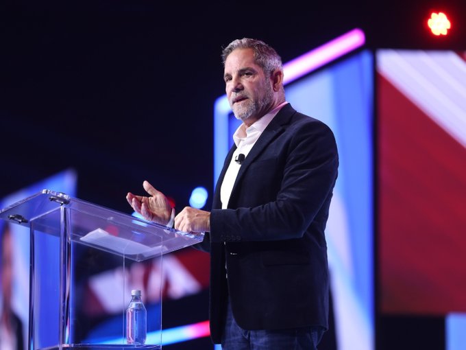 The image features a man standing at a podium with his hands outstretched, giving a speech. The background of the image shows a stage with a large screen behind him, which displays a message from Grant Cardone. The text on the screen reads "LATEST: Grant Cardone says his wealth strategy is to hustle fiat and convert it to real estate or Bitcoin, saying he doesn't believe in gold and silver." This suggests that the man speaking at the podium might be discussing financial matters or sharing insights about investing strategies.