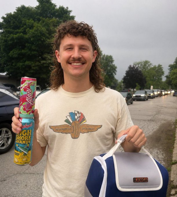 The image shows a man with long hair standing next to a bag, holding a can of soda or energy drink. The photo has been edited to include text that makes him look like an older person, creating a humorous effect as if he's mimicking his favorite actor or character from an older movie. The context behind the edited image is unclear, but it seems to be related to the post about him being an artist or someone who loves Mullet movies. The main focus of the photo remains the man and the can of soda in his hand.