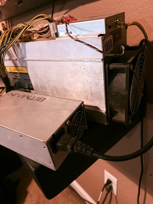 A Bitmain Bitcoin mining rig (Antminer, serial 1898) with power supplies and cooling fans set up on a shelf, plugged into a wall outlet.