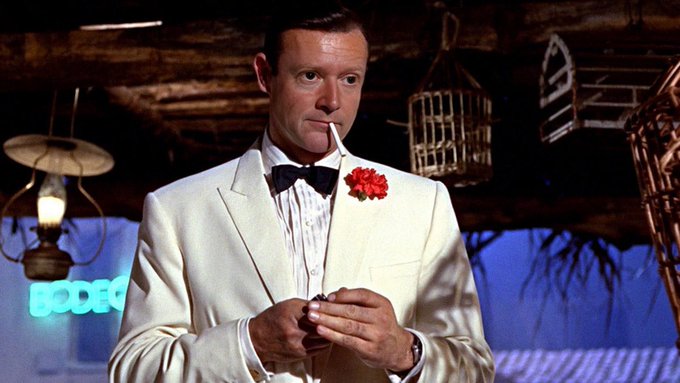 A photograph of a man in a white tuxedo with a black bowtie and red carnation, smoking a cigarette, in a rustic setting with a birdcage and "BEDEC" neon sign visible in the background.