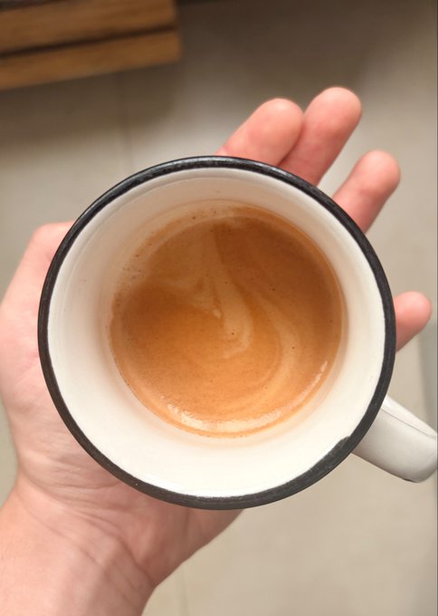 A close-up view looking down into a white mug with a black rim, held in a hand, containing a light brown coffee beverage with foam.