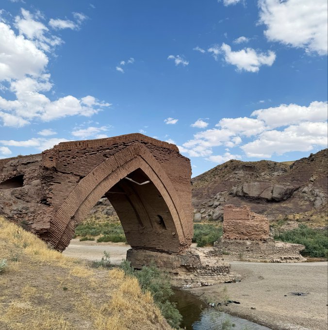 An old brick arch bridge in a desert landscape under a blue sky with scattered clouds, with a dry riverbed below and hills in the background.