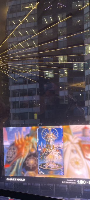 A nighttime photograph showing a digital billboard advertising the mobile game 'GHAZZ GOLD' (by J.A. Mobile, rated 100+) with religious/spiritual imagery, reflected on a building facade with yellow string lights.