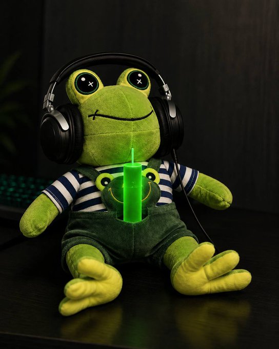 A green frog plush toy wearing black headphones and overalls sits on a dark surface, holding a glowing green lightsaber, perfectly matching the "$FROG" crypto ticker theme in the post.