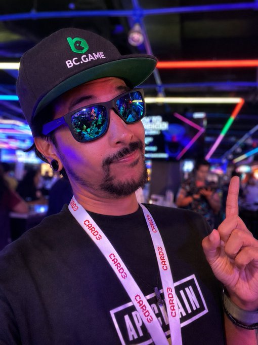 A man poses at what appears to be a crypto or gaming conference, wearing a "BC.GAME" cap and a "CARD3" lanyard while pointing upward. His reflective sunglasses show flashes of the colorful event lighting, matching the casual "Gn fam" (good night family) sign-off vibe of a content creator wrapping up their day at the event.