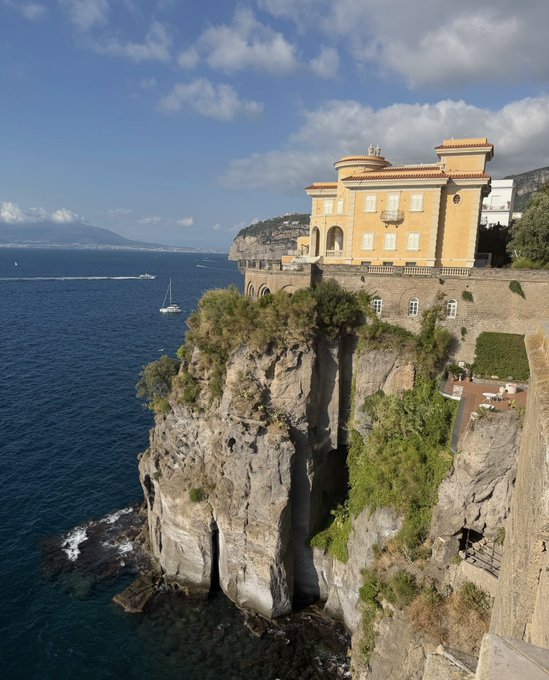 An aerial view of a yellow Mediterranean-style villa perched atop a steep rocky cliff overlooking a deep blue sea, with sailboats visible in the distance.
