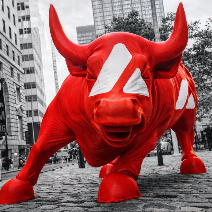 A striking red inflatable bull statue standing on a cobblestone street with a desaturated urban cityscape in the background, featuring a stylized white logo on its forehead and side.
