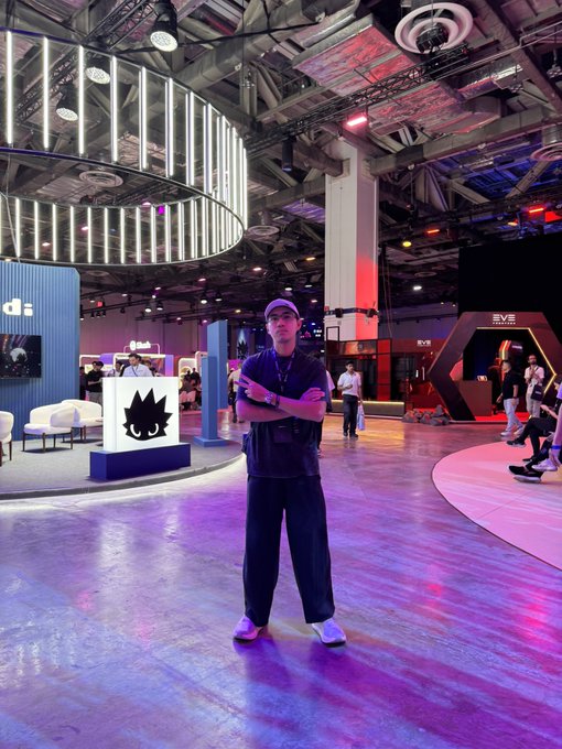 A young man in a black shirt, cap, and gold chain stands confidently with crossed arms in a vibrant convention hall illuminated by purple and red lighting, with "EVE Frontier" signage visible in the background. The image reflects the energetic "SUI Basecamp aura" of the @SuiNetwork $SUI event.