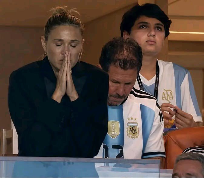 A woman in a black blazer prays with her hands clasped beside a man and boy wearing Argentina (AFA) football jerseys, appearing somber as they watch a match.