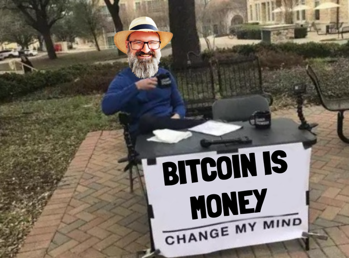 A bearded man wearing a straw hat and glasses sits at a table with a 'BITCOIN IS MONEY CHANGE MY MIND' sign, holding a coffee mug in an outdoor setting.