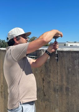 A person wearing a cap, sunglasses, and a tan shirt working with tools on a concrete wall outdoors on a sunny day.