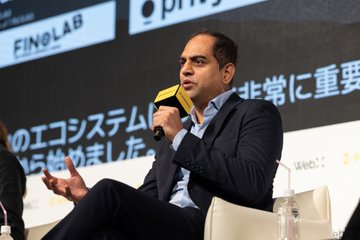 A man in a dark suit speaks into a yellow microphone while seated on a white chair at a panel event, with Japanese text visible on the screen behind him including 'FINOLAB' and 'privy' logos.