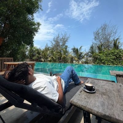 A person relaxing on a lounge chair by a swimming pool with tropical trees, holding a drink, with a cup of coffee on the side table.