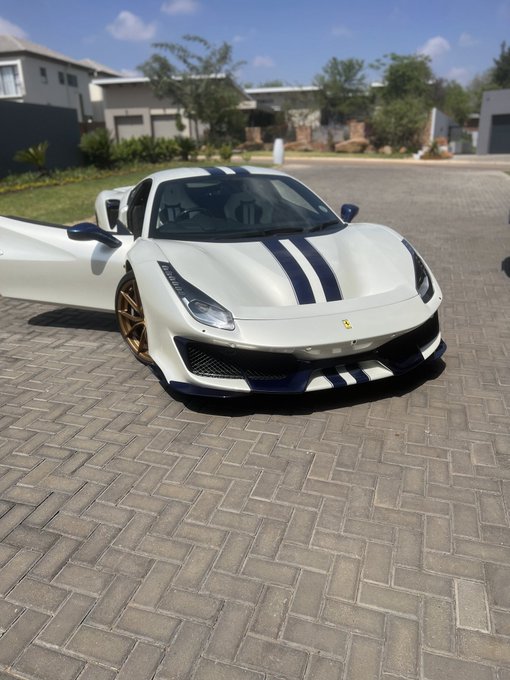 A white Ferrari sports car with blue racing stripes and gold wheels parked on a paved driveway in front of modern houses.