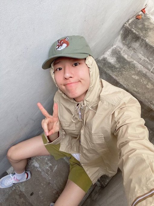 A selfie of a young woman wearing a beige hooded jacket, green shorts, and a green cap with a fox patch, making a peace sign while sitting on concrete steps.