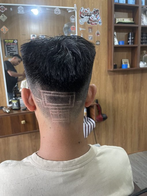 The back of a person's head showing a fresh haircut with a faded design cut into the nape, taken in a barbershop with wooden shelves and decorations visible in the background.