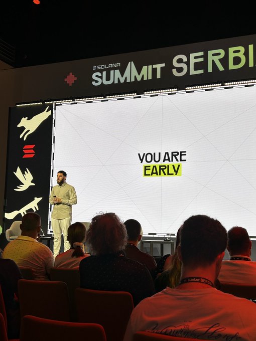 A speaker on stage at the 'SOLANA SUMMIT SERBIA' addressing an audience, with the words 'YOU ARE EARLY' displayed on a large screen behind him.