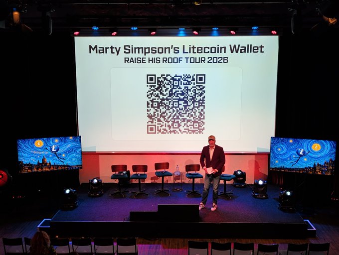 The image shows a stage with a man standing at the front of the room, giving a speech or presentation. The stage has a large screen behind him displaying a QR code that reads "Marty'S @nexuswallet LTC address, hitting up the crowd for Litecoin". This QR code is likely related to the event being held on this stage and serves as an invitation for attendees to learn more about Litecoin or participate in a fundraising event.
