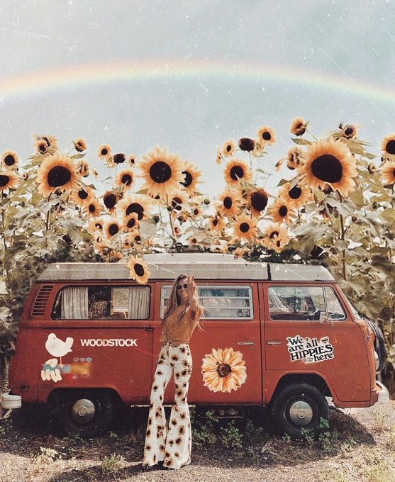 A vintage-style retro photograph of a woman in 1970s flared pants standing in front of a red VW bus with 'WOODSTOCK' and 'We are all hippies here' decals, set against a sunflower field and a rainbow sky.