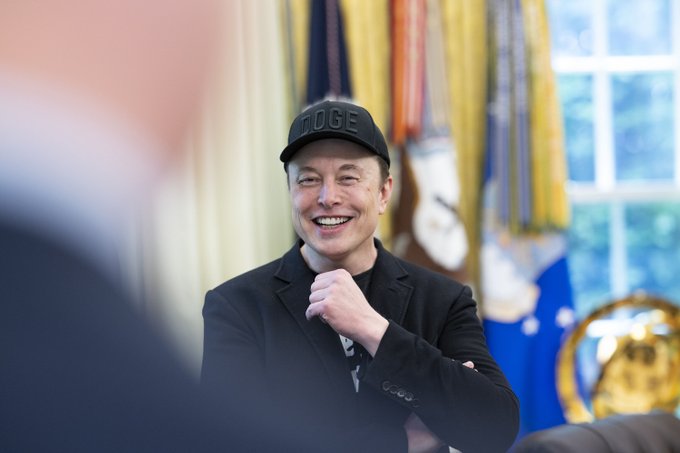 Elon Musk smiles while wearing a black "DOGE" cap inside what appears to be the White House Oval Office. The image relates to the post because Musk's SpaceX joining the Nasdaq-100 adds to his presence in major market indexes, with his companies collectively holding significant Bitcoin reserves.
