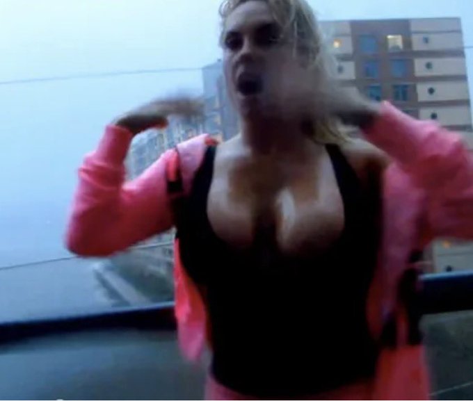 A blonde woman wearing sunglasses and a pink jacket over a black top is dancing or moving energetically on what appears to be a balcony.