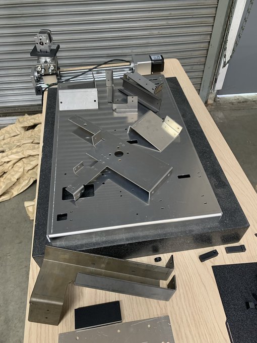 A metal fabrication project showing various cut and bent steel pieces arranged on a granite surface plate on a wooden workbench, with what appears to be a bandsaw in the background in a workshop setting.