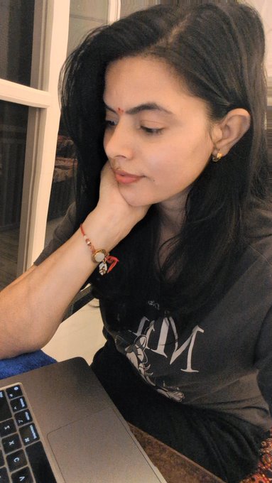 A young woman with dark hair tied back, wearing a black graphic t-shirt and a bindi, rests her head on her hand while looking thoughtfully at a laptop screen.