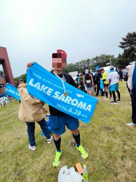 The image shows a runner proudly holding a "LAKE SAROMA 100km" sign with the date "2018.6.24 SUN" after completing the Saroma Lake 100km Ultramarathon in Japan, celebrating their achievement with another finisher.