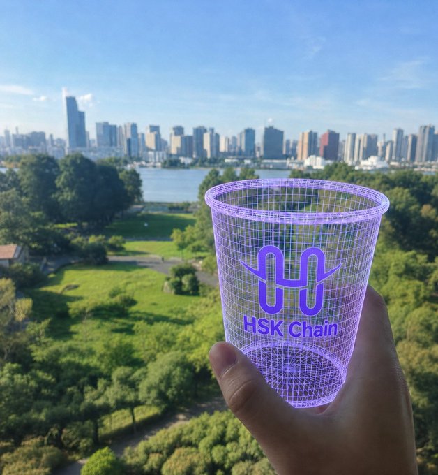 A hand holds up a wireframe holographic cup branded "HSK Chain" with a stylized "H" logo against a real-world city skyline and green park, visually blending the physical landscape with a digital blockchain overlay that reflects the post's message of an "on-chain layer" on top of reality.