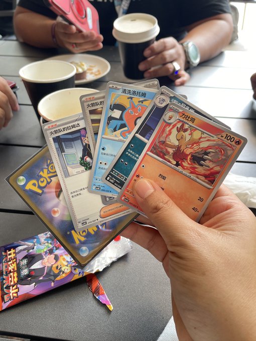 A hand holding a fan of Chinese Pokémon trading cards featuring characters like Cinderace (炽焰咆哮虎) and Battle Coin cards at an outdoor table with coffee cups.