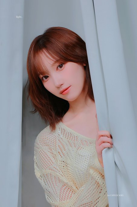 A portrait of a young woman with short brown hair peeking through light blue curtains, wearing a cream-colored knit top with the text 'lights...' and 'lights IN NEW SOO'.