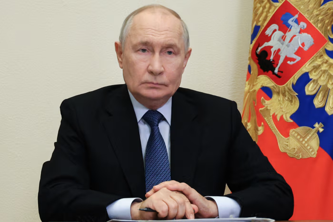 Vladimir Putin sits with hands clasped in front of the Russian flag bearing the double-headed eagle coat of arms.