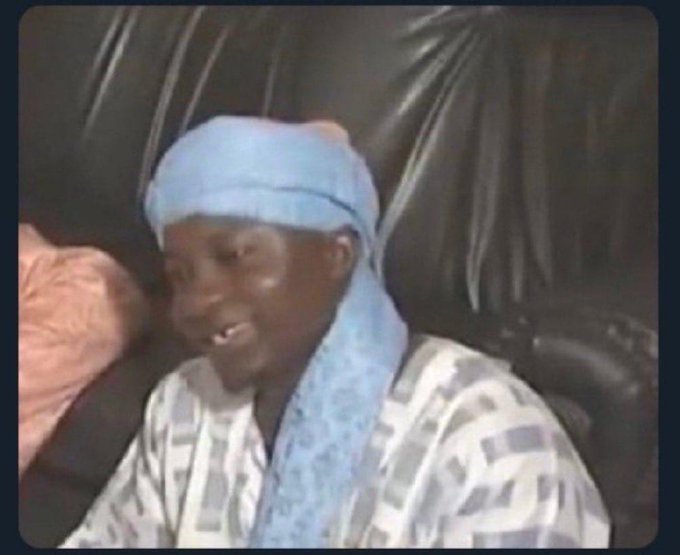 Screenshot from a video showing a young boy wearing a light blue head covering and a patterned shirt, smiling while sitting on a black leather couch.