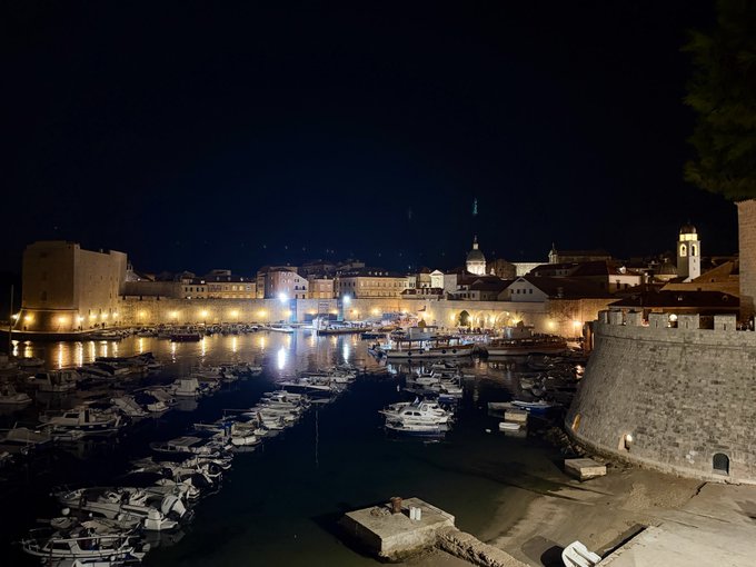 Nighttime view of a harbor filled with small boats, surrounded by illuminated stone buildings and towers.