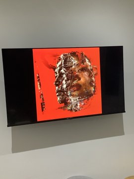 A framed painting with a bright orange background depicts a skull-like, decaying face with metallic, textured details and dark text along the left side.