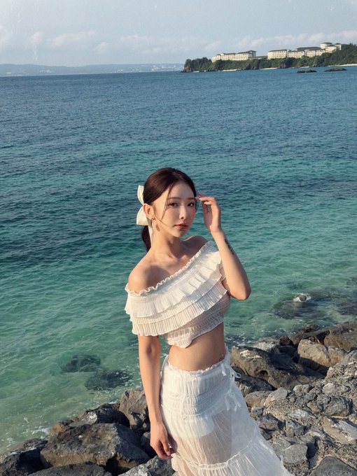 A young woman in a white off-shoulder ruffled crop top and matching skirt stands on rocky shoreline with turquoise ocean water and a coastline with buildings visible in the background.
