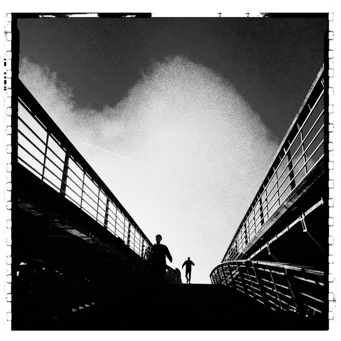 The image shows two people walking up a bridge, one of them holding a suitcase. The scene appears to be taken during daylight hours with an overcast sky. This setting evokes a sense of contrast between the natural light coming from the sky and the darkness below the bridge as well as the urban landscape surrounding it. The two figures' presence adds an element of tension, as they navigate the stairs up the structure, possibly indicating that they are travelers or adventurers exploring the cityscape.