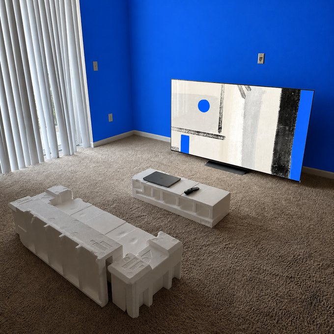 A blue-walled living room features a couch and coffee table sculpted entirely from styrofoam, alongside a flat-screen TV displaying abstract art. The meme humorously critiques crypto culture by depicting minimalist, makeshift furniture as the ultimate lifestyle goal for crypto enthusiasts aiming for eight-figure success.