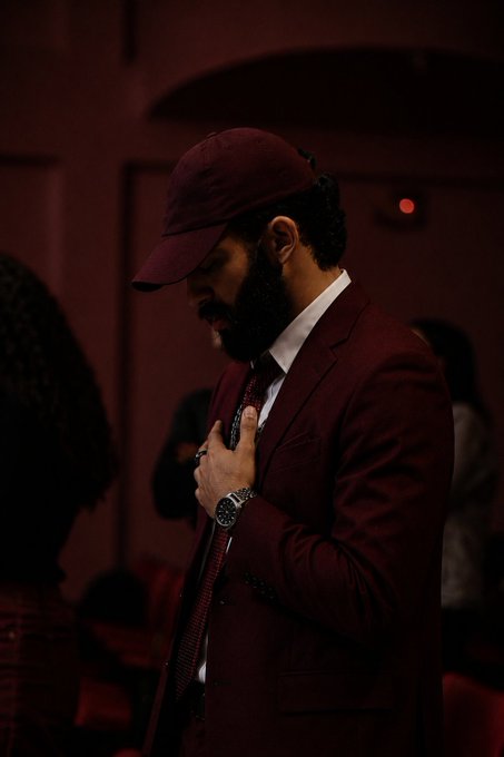 A man in a maroon suit, cap, and tie with a beard is photographed in a dimly lit room with red lighting.