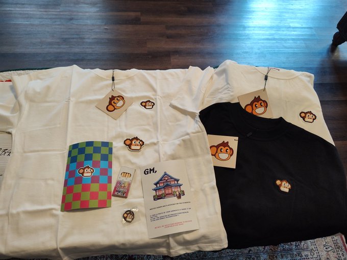 A flat-lay photo of two t-shirts (one white, one black) with monkey face tags, a card reading 'GM,', a colorful checkered card, and pins on a wooden floor.