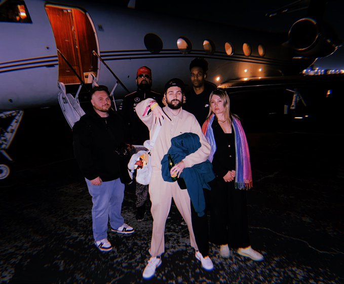 A group of five people pose at night in front of a private jet with illuminated cabin windows and a deployed airstair. The central figure wears a matching cream-colored outfit while holding a drink, and the luxurious travel scene reflects the post's theme of enjoying a high-flying lifestyle after ANSEM's $PUMP trade flip.