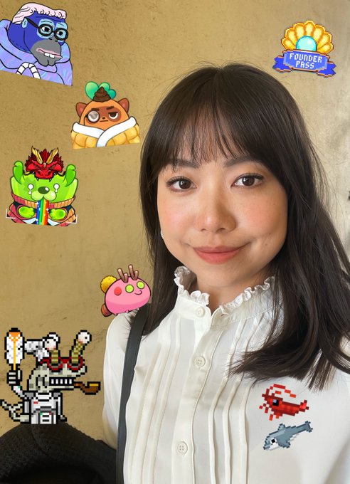 The image features a woman with long brown hair wearing a white shirt, posing for a selfie against a wall adorned with various cartoon characters and logos. The woman's face is lit up with a smile as she looks directly at the camera. The background includes a cartoon character, a pink fish, and other cartoon-themed elements that add to the overall playful atmosphere of the image.