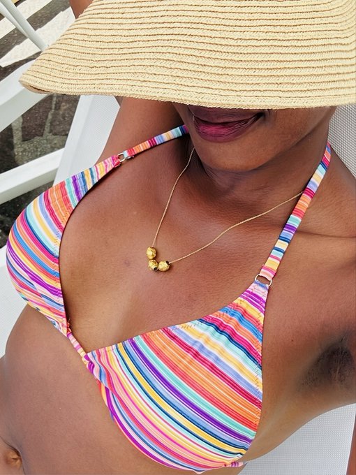 A close-up photo of a woman wearing a rainbow-striped bikini, a straw sun hat, and a gold necklace with small charms.