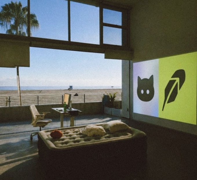 An atmospheric room overlooking a beach with a mattress on the floor, a desk with a monitor, and a large projection screen displaying the GitHub and Atom logos.