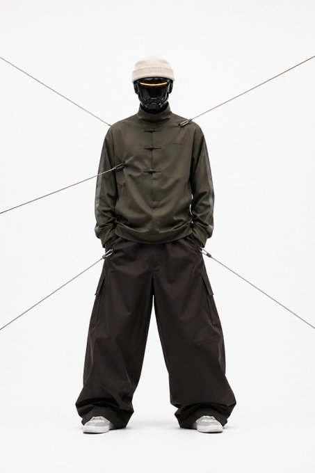 A full-body studio shot of a person wearing techwear streetwear: an olive green pullover jacket, dark brown cargo pants, a beanie, and a black face mask, standing against a white background with diagonal lines.