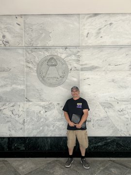 A man wearing a black graphic t-shirt and a cap stands in front of a marble wall featuring an embossed seal, posing for a photo to advocate for the CLARITY Act during a visit to Senator Wyden's Portland field office alongside PNW Block representatives.
