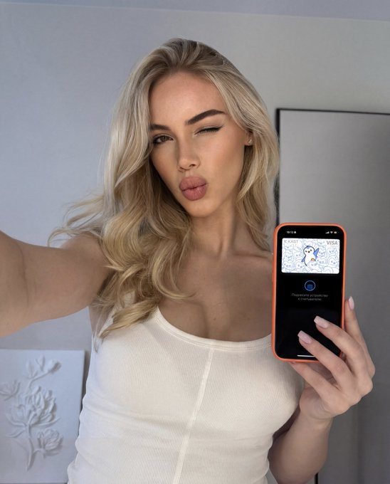 The image features a woman with blonde hair, wearing a white tank top, holding up her cell phone to take a selfie. The photo includes text that reads "GM fam Now’s the perfect time to get your KAST & Pudgy Penguins Card and start taking advantage of all its benefits". This suggests that the post is promoting a credit card company called Kast & Pudgy, which offers rewards for purchases made at participating retailers. The woman's selfie with her cell phone serves as an advertisement for this credit card company.
