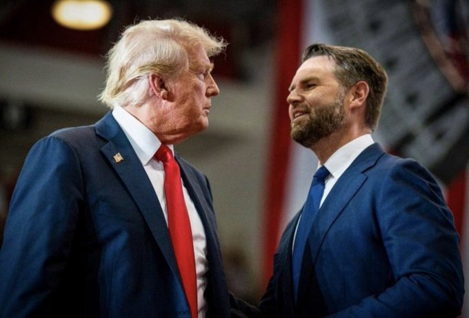 President Donald Trump, in a dark suit with a red tie, stands face-to-face with Vice President JD Vance, wearing a navy suit and blue tie, appearing to share a smile during an in-person exchange. The image visually accompanies the milestone announcement that both the President and Vice President now officially hold Bitcoin.