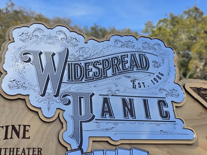 A custom wooden sign featuring ornate lettering that reads "Widespread Panic Est. 1986" is displayed outdoors against a backdrop of trees and blue sky, likely commemorating a piece of memorabilia from the long-running jam band. The "Got one!!" caption suggests the photographer is proud of acquiring this collectible concert merchandise.