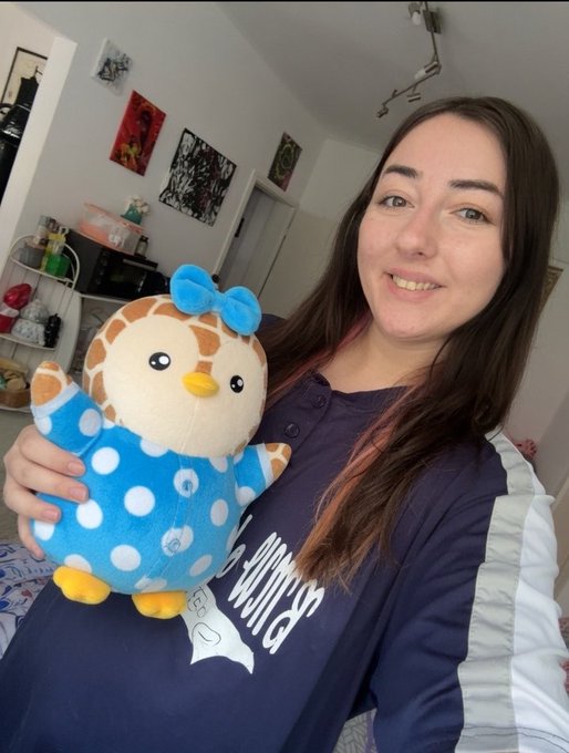 A selfie of a smiling woman with long brown hair holding a cute plush penguin toy wearing a blue polka dot outfit and a blue bow, taken in a room with artwork on the walls.